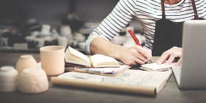 A woman taking notes at in her ceramics studio as she looks at her open laptop.