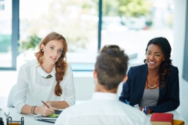 Two business women sit at a desk and talk across the desk to a man.